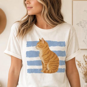 Könnte beinhalten: Ein cremefarbenes Kurzarm-T-Shirt mit einer Grafik einer orangefarbenen getigerten Katze, die im Profil sitzt. Die Katze ist über vier horizontalen, abgenutzten blauen Streifen in der Mitte des T-Shirts platziert. Das T-Shirt hat einen Rundhalsausschnitt.
