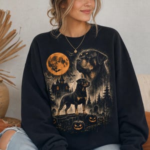 Könnte beinhalten: Ein schwarzes Langarm-Sweatshirt mit einem Grafikdruck eines Rottweilers, einem großen orangefarbenen Vollmond, einem Spukhaus, Tannenbäumen und Kürbislaternen. Das Design verwendet Orange-, Gelb- und Schwarztöne für eine gruselige Szene.