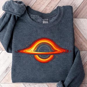 May include: A dark gray sweatshirt featuring a colorful embroidered design of a black hole. The design includes orange, yellow, and red hues, with a circular center and radiating lines. The sweatshirt is folded on a wooden surface.
