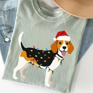 Puede incluir: Camiseta verde claro con un beagle de dibujos animados con un gorro de Papá Noel y luces navideñas. El perro tiene marcas negras, blancas y marrones. Un sombrero color canela y unos vaqueros azules están en el fondo.