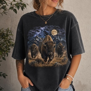 May include: A charcoal grey short-sleeve t-shirt featuring a graphic print of three bison running across a grassy plain. The background depicts a dark, stormy sky with lightning bolts and a full moon above mountain peaks in the distance.