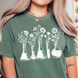 Può includere: Una maglietta verde con una grafica bianca di fiori in becher. La grafica è centrata sulla maglietta.