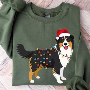 Peut inclure: Sweat-shirt vert avec un dessin de chien portant un bonnet de Père Noël et des guirlandes lumineuses. Le chien est noir, blanc et marron. Le sweat-shirt est de couleur verte unie. Les manches sont retroussées.