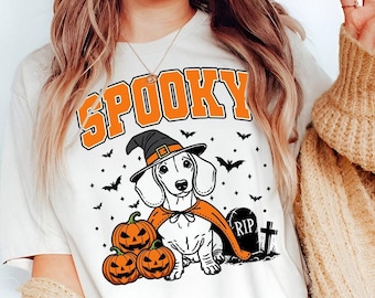 Op de afbeelding: Wit T-shirt met een afbeelding van een teckel die een heksenhoed en een cape draagt en een pompoen vasthoudt. De tekst "SPOOKY" staat boven de hond. Er zijn vleermuizen en sterren op de achtergrond.