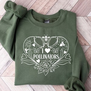Puede incluir: Una sudadera verde con un gráfico blanco en forma de corazón con el texto "Me encantan los polinizadores" en su interior. El corazón está rodeado de flores, abejas, mariposas y otros insectos.