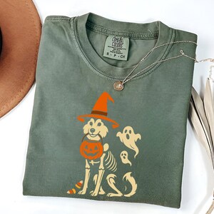 May include: Sage green t-shirt featuring a Halloween-themed graphic. The design includes a dog wearing a witch hat and holding a pumpkin bucket, with a skeleton outline and two ghost figures. A silver necklace and a brown hat are also visible.