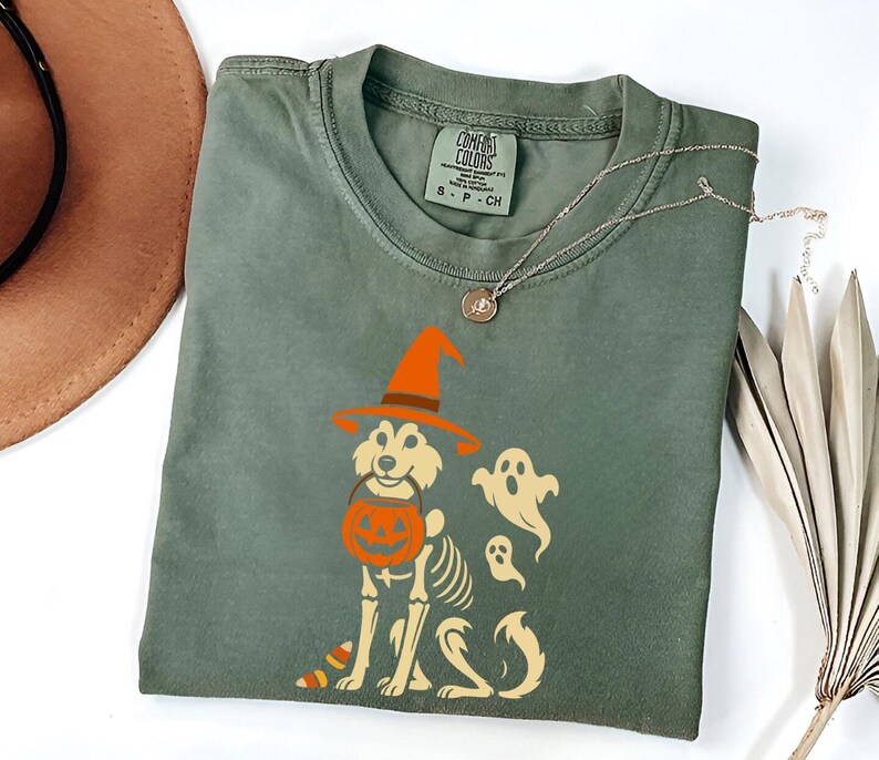 May include: Sage green t-shirt featuring a Halloween-themed graphic. The design includes a dog wearing a witch hat and holding a pumpkin bucket, with a skeleton outline and two ghost figures. A silver necklace and a brown hat are also visible.