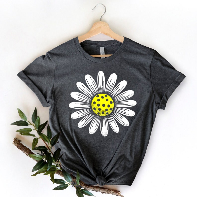 May include: A dark gray t-shirt with a white daisy flower graphic. The center of the flower is a yellow and black pickleball.