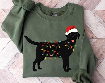 Può includere: Felpa verde con un'illustrazione di un labrador nero con un cappello da Babbo Natale e luci natalizie. Il cane ha la lingua di fuori. La felpa è di colore verde unito.