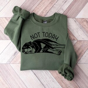May include: An olive green sweatshirt with the words "NOT TODAY" above a black and white graphic of a dog lying down. The sweatshirt has long sleeves with ribbed cuffs.