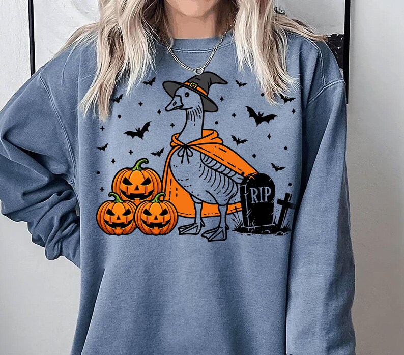 May include: A blue sweatshirt with a graphic of a goose wearing a witch hat and holding a pumpkin. The goose is standing in front of a tombstone that says "RIP". There are three pumpkins in the bottom left corner of the image.