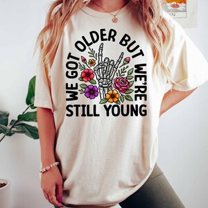 May include: A white t-shirt with a black circle graphic featuring a skeleton hand making a rock and roll sign surrounded by colorful flowers. The text "We got older but we're still young" is written around the graphic.