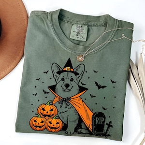 May include: A green t-shirt with a black and orange illustration of a corgi wearing a witch's hat and cape. The corgi is surrounded by bats, stars, and pumpkins. The text "RIP" is on a tombstone.