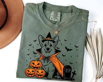 May include: A green t-shirt with a black and orange illustration of a corgi wearing a witch's hat and cape. The corgi is surrounded by bats, stars, and pumpkins. The text "RIP" is on a tombstone.