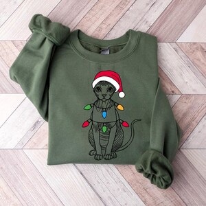 May include: A green sweatshirt with a black and white illustration of a cat wearing a red Santa hat and Christmas lights.