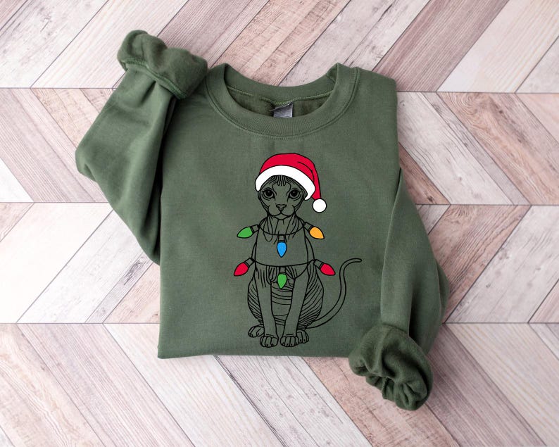 May include: A green sweatshirt with a black and white illustration of a cat wearing a red Santa hat and Christmas lights.