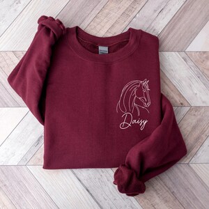 May include: Burgundy sweatshirt with a white outline of a horse's head and the name "Daisy" embroidered on the front.