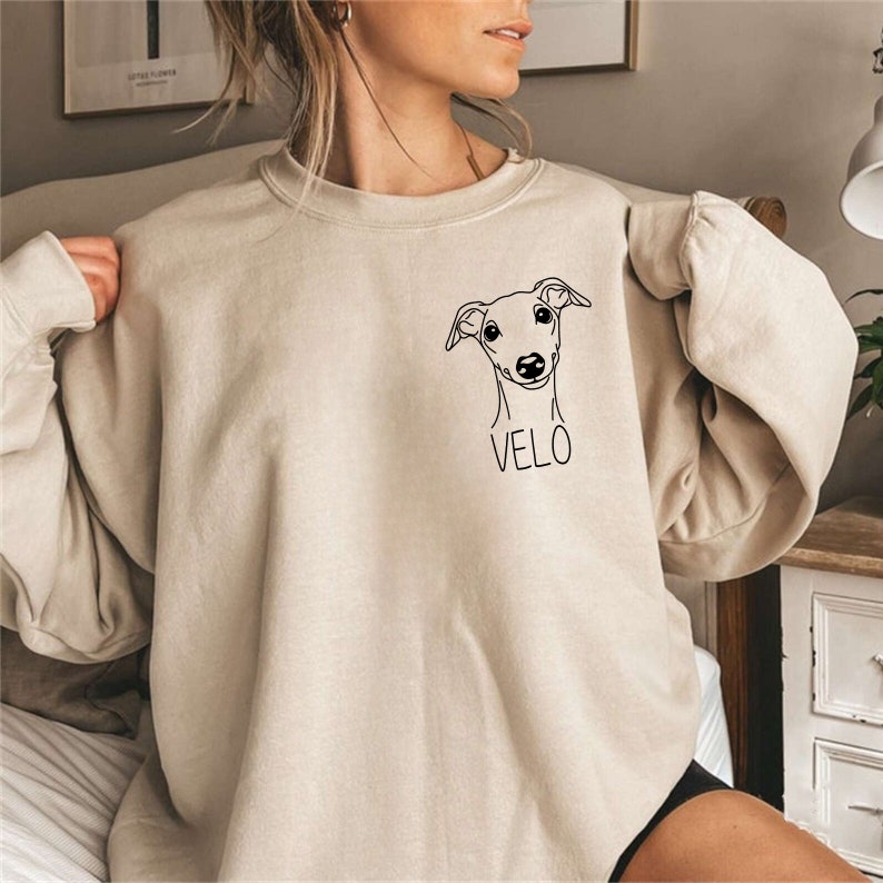 K&ouml;nnte beinhalten: Ein beiger Pullover mit einer schwarzen Umrandung eines Hundekopfes und dem darunter gedruckten Namen "VELO".
