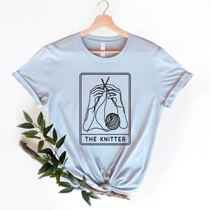 May include: A light blue t-shirt featuring a black line drawing of a hand knitting with the text "THE KNITTER" beneath the illustration.