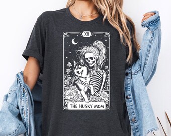 May include: A dark gray t-shirt with a white graphic of a skeleton holding a husky dog. The graphic is in the style of a tarot card and says "The Husky Mom" at the bottom.