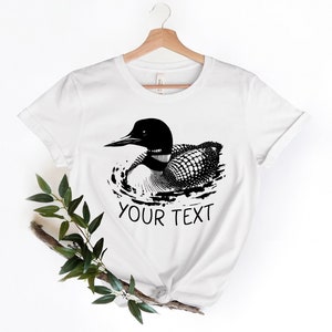 May include: White t-shirt with a black and white illustration of a loon swimming. The text "YOUR TEXT" is printed below the loon.