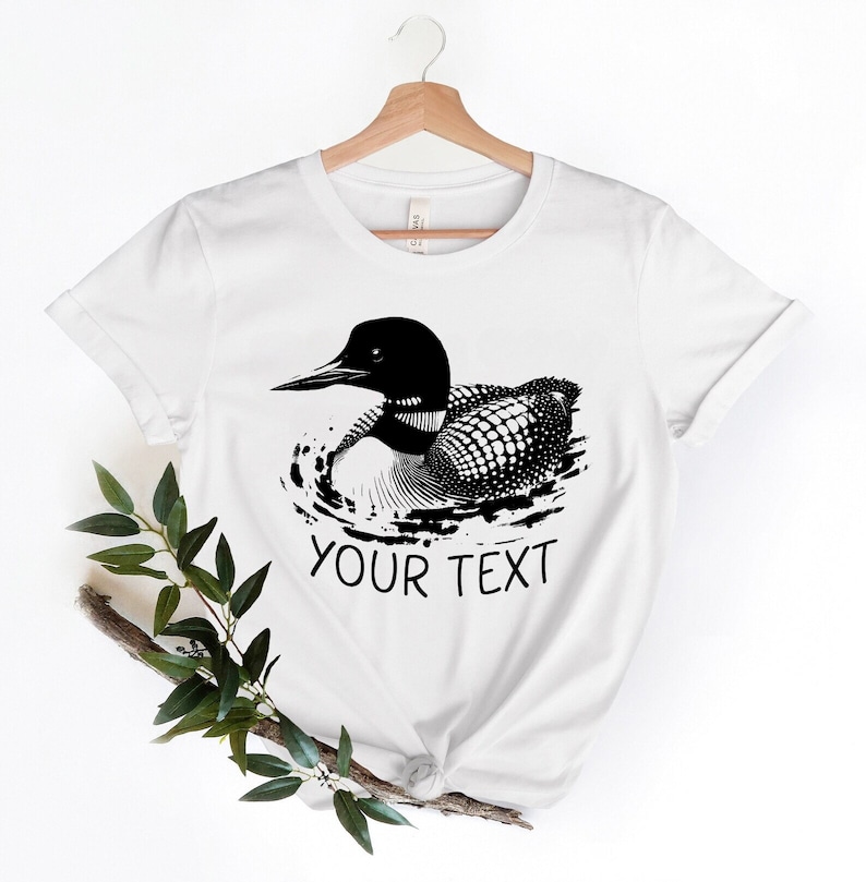 May include: White t-shirt with a black and white illustration of a loon swimming. The text "YOUR TEXT" is printed below the loon.