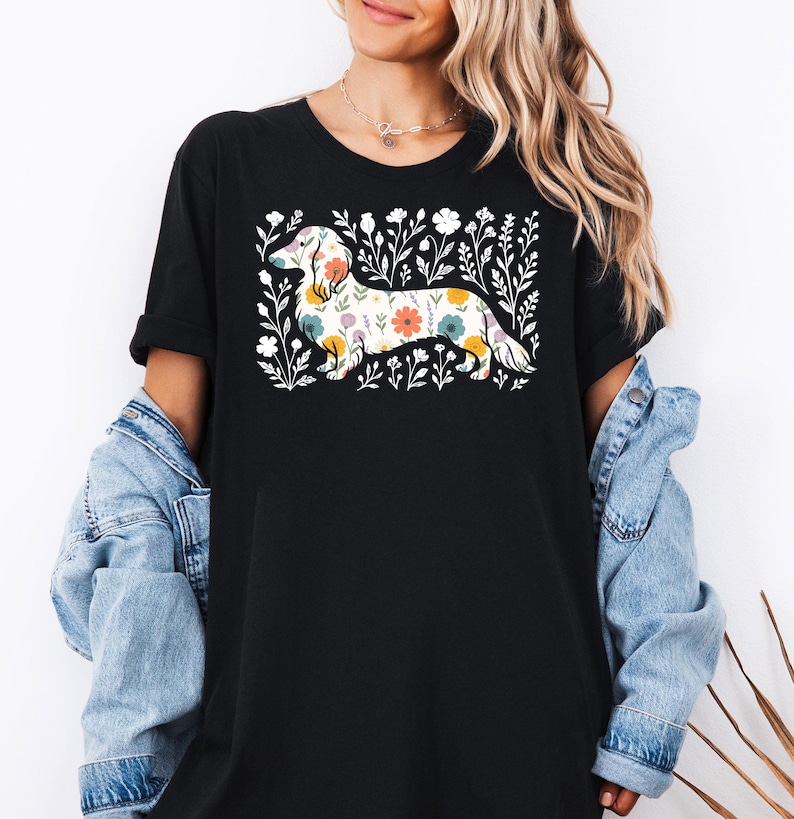 May include: Black t-shirt featuring a floral dachshund design. The dachshund is filled with colorful flowers and surrounded by white floral accents. The shirt has short sleeves and a relaxed fit.