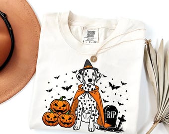 May include: A white t-shirt with a black and white dalmatian dog wearing a witch's hat and orange cape. The dog is surrounded by pumpkins and bats. The text "RIP" is on a tombstone.