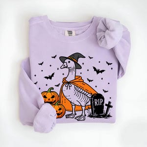 May include: A lavender sweatshirt with a graphic of a goose wearing a witch hat and holding a pumpkin. The goose is standing next to a tombstone that says "RIP". There are bats and pumpkins scattered around the goose.
