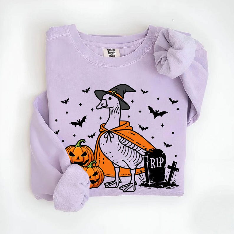 May include: A lavender sweatshirt with a graphic of a goose wearing a witch hat and holding a pumpkin. The goose is standing next to a tombstone that says "RIP". There are bats and pumpkins scattered around the goose.