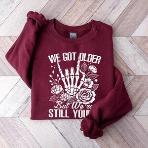 May include: Burgundy sweatshirt with a white graphic of a skeleton hand giving the rock and roll sign with flowers. The text reads "We got older but we're still you".