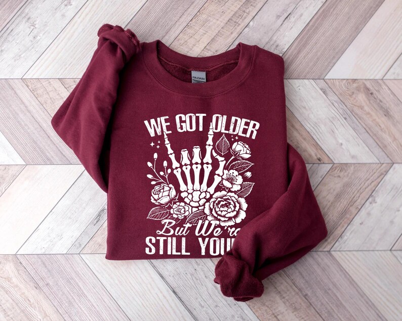 May include: Burgundy sweatshirt with a white graphic of a skeleton hand giving the rock and roll sign with flowers. The text reads "We got older but we're still you".