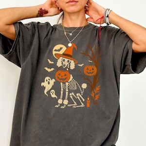 May include: Dark grey t-shirt with a Halloween design. The graphic depicts a poodle skeleton wearing a witch's hat and holding a jack-o'-lantern. Other elements include bats, ghosts, a full moon, and a tree with a pumpkin.