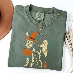 May include: Sage green Comfort Colors t-shirt featuring a Halloween-themed graphic. The design includes a dog wearing a witch hat and carrying a pumpkin, a ghost, bats, a crescent moon, and a candle. A silver necklace is draped across the shirt.