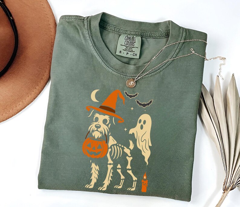May include: Sage green Comfort Colors t-shirt featuring a Halloween-themed graphic. The design includes a dog wearing a witch hat and carrying a pumpkin, a ghost, bats, a crescent moon, and a candle. A silver necklace is draped across the shirt.