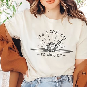 May include: A white t-shirt with a black graphic of a yarn ball and knitting needles with the text "It's a good day to crochet".