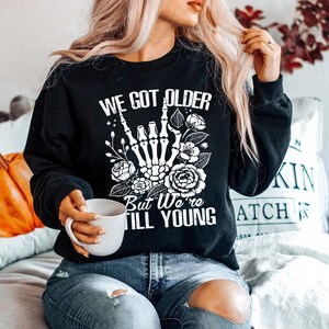 May include: Black sweatshirt with a white graphic print that says "We got older but we're still young". The graphic features a skeleton hand giving the rock and roll sign with flowers around it.