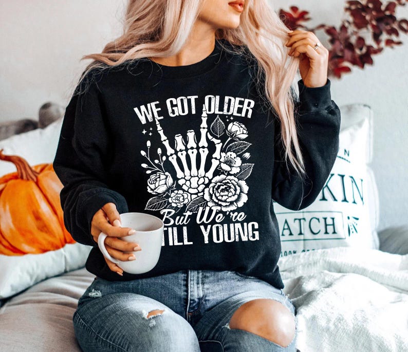 May include: Black sweatshirt with a white graphic print that says "We got older but we're still young". The graphic features a skeleton hand giving the rock and roll sign with flowers around it.