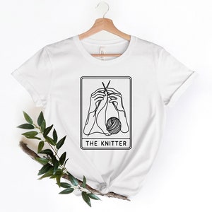 K&ouml;nnte beinhalten: Wei&szlig;es T-Shirt mit einer schwarzen Strichzeichnung von H&auml;nden, die mit Nadeln und Garn stricken. Das Bild befindet sich in einem rechteckigen Rahmen mit dem Text "THE KNITTER" darunter. Das T-Shirt h&auml;ngt an einem Holzb&uuml;gel.