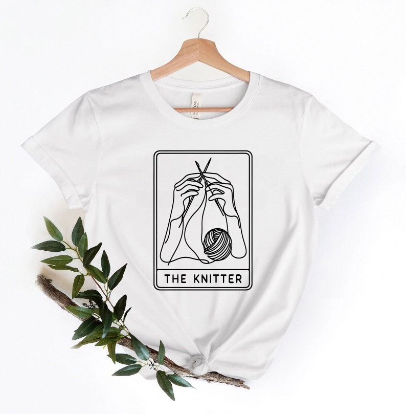 K&ouml;nnte beinhalten: Wei&szlig;es T-Shirt mit einer schwarzen Strichzeichnung von H&auml;nden, die mit Nadeln und Garn stricken. Das Bild befindet sich in einem rechteckigen Rahmen mit dem Text "THE KNITTER" darunter. Das T-Shirt h&auml;ngt an einem Holzb&uuml;gel.