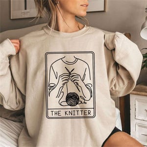 A knitting sweatshirt with a black graphic print of a person knitting with the text 'THE KNITTER' below the graphic.