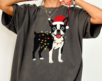 Puede incluir: Camiseta gris oscuro con un perro Boston Terrier con un gorro de Papá Noel y luces de cadena. El perro es blanco y negro con la lengua roja. Las luces de cadena son multicolores. Un diseño festivo de vacaciones.