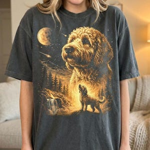 May include: Dark gray t-shirt featuring a detailed illustration of a golden doodle dog with a night sky backdrop. The design includes a full moon, stars, and a smaller dog silhouette. The shirt has a relaxed fit.