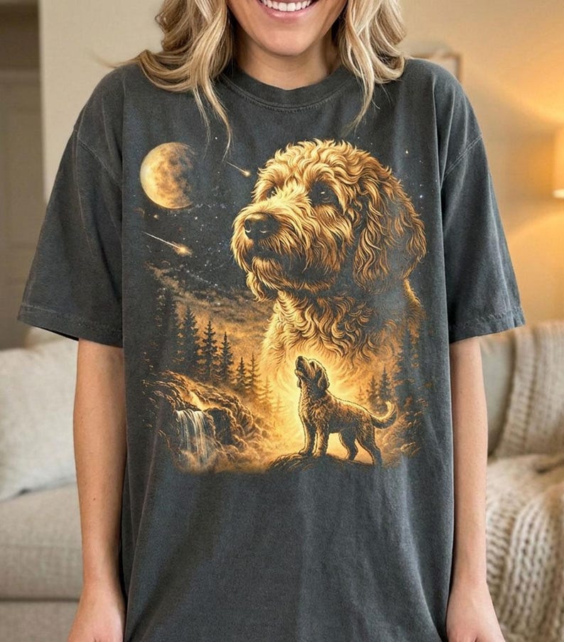 May include: Dark gray t-shirt featuring a detailed illustration of a golden doodle dog with a night sky backdrop. The design includes a full moon, stars, and a smaller dog silhouette. The shirt has a relaxed fit.