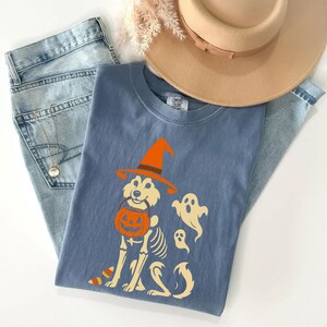 May include: A blue Comfort Colors t-shirt featuring a Halloween-themed graphic of a dog skeleton wearing a witch hat and holding a jack-o'-lantern. Two ghost figures are also present. The shirt is paired with light-wash jeans and a tan hat.
