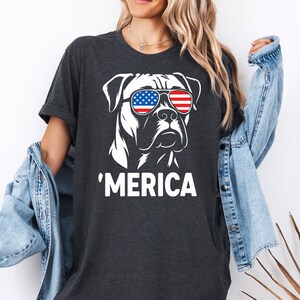 May include: Dark grey t-shirt with a white graphic of a dog wearing sunglasses in the style of the American flag. The word "'MERICA" is printed in white below the dog illustration. The shirt has short sleeves.