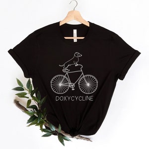 May include: Black t-shirt with a white outline of a dachshund riding a bicycle. The text "DOXYCYCLINE" is printed below the bicycle.