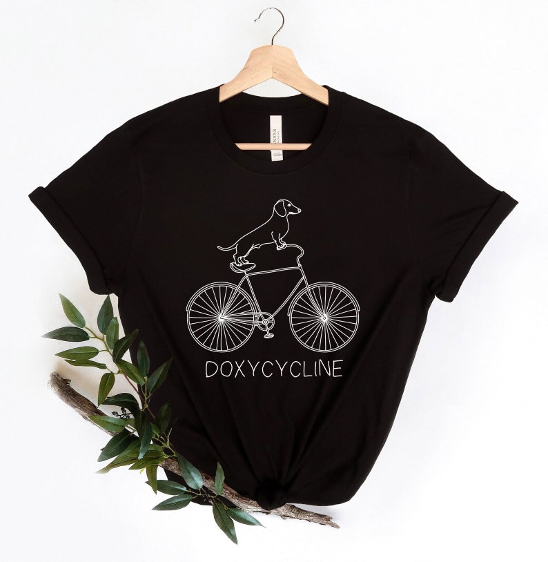 May include: Black t-shirt with a white outline of a dachshund riding a bicycle. The text "DOXYCYCLINE" is printed below the bicycle.