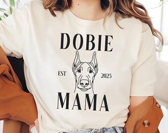 May include: White t-shirt with black text that reads "Dobie Est 2025 Mama" and a line drawing of a Doberman Pinscher.