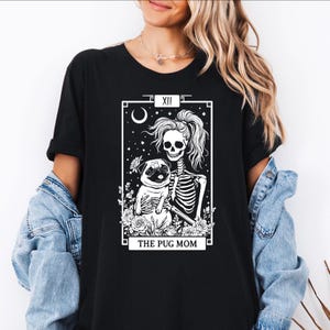 Może przedstawiać: Czarny t-shirt z białym nadrukiem przedstawiającym szkielet trzymający mopsa, z napisem "THE PUG MOM". Projekt jest oprawiony w prostokątną ramkę z księżycem i gwiazdami.